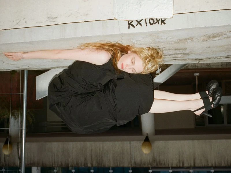 A young woman with red hair and wearing a black dress poses in an unusual pose and wearing high heels in a low light setting.