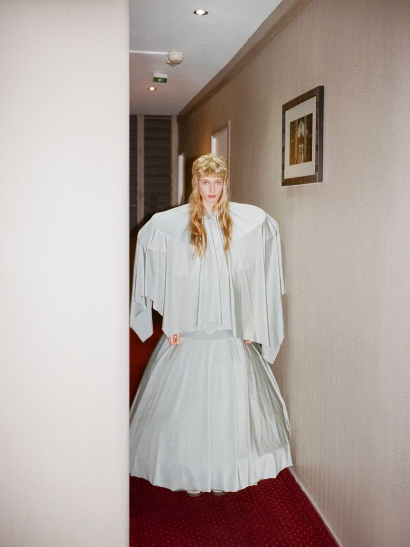 A fair-skinned person with blonde hair poses in an oversized light blue dress in a hallway with pale beige walls and a red patterned carpet.