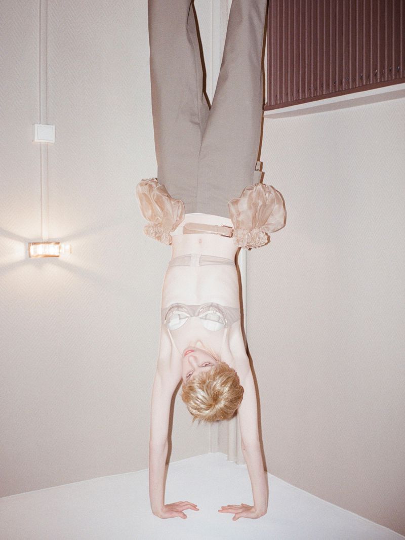 Adult female with short blonde hair does a handstand in lingerie and trousers in an indoor corner studio with minimalist lighting.