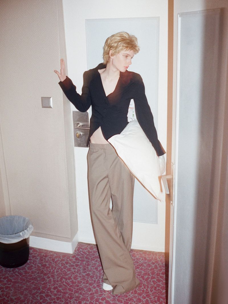 A fair-skinned, blonde-haired woman in a black top and khaki pants poses with a white pillow in an indoor setting with patterned carpet.
