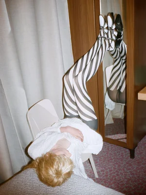 Model lying upside down on a white chair with legs raised wearing striped tights and white shoes against a hotel room background