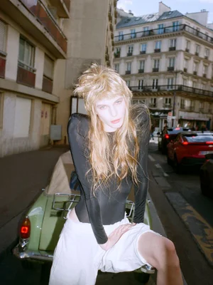 Model with long blonde hair in a black long-sleeve top and white shorts leaning on a vintage car on a city street.