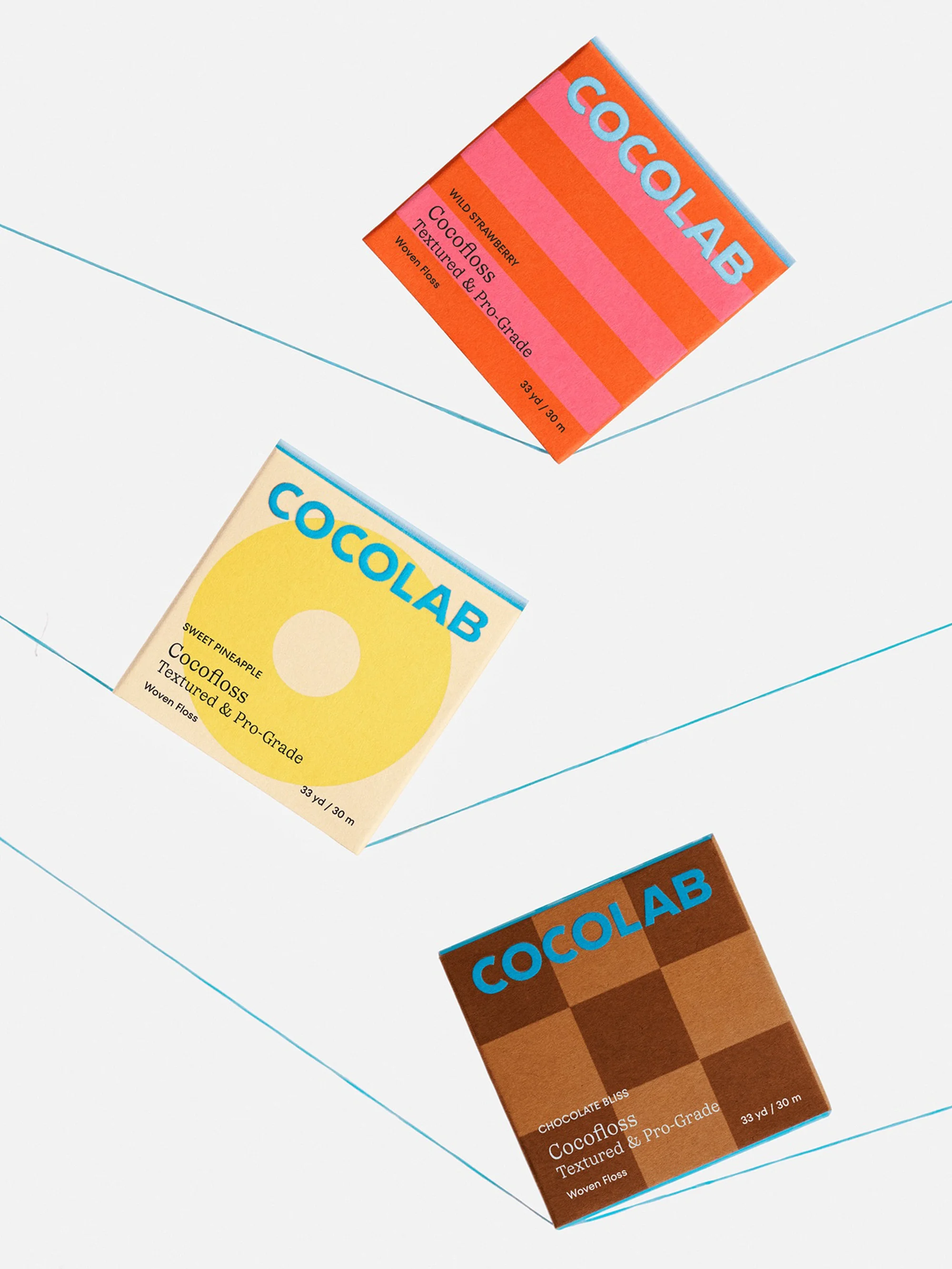 Three Cocofloss dental floss packages are displayed in a studio setting. Each package has a different color and flavor theme.