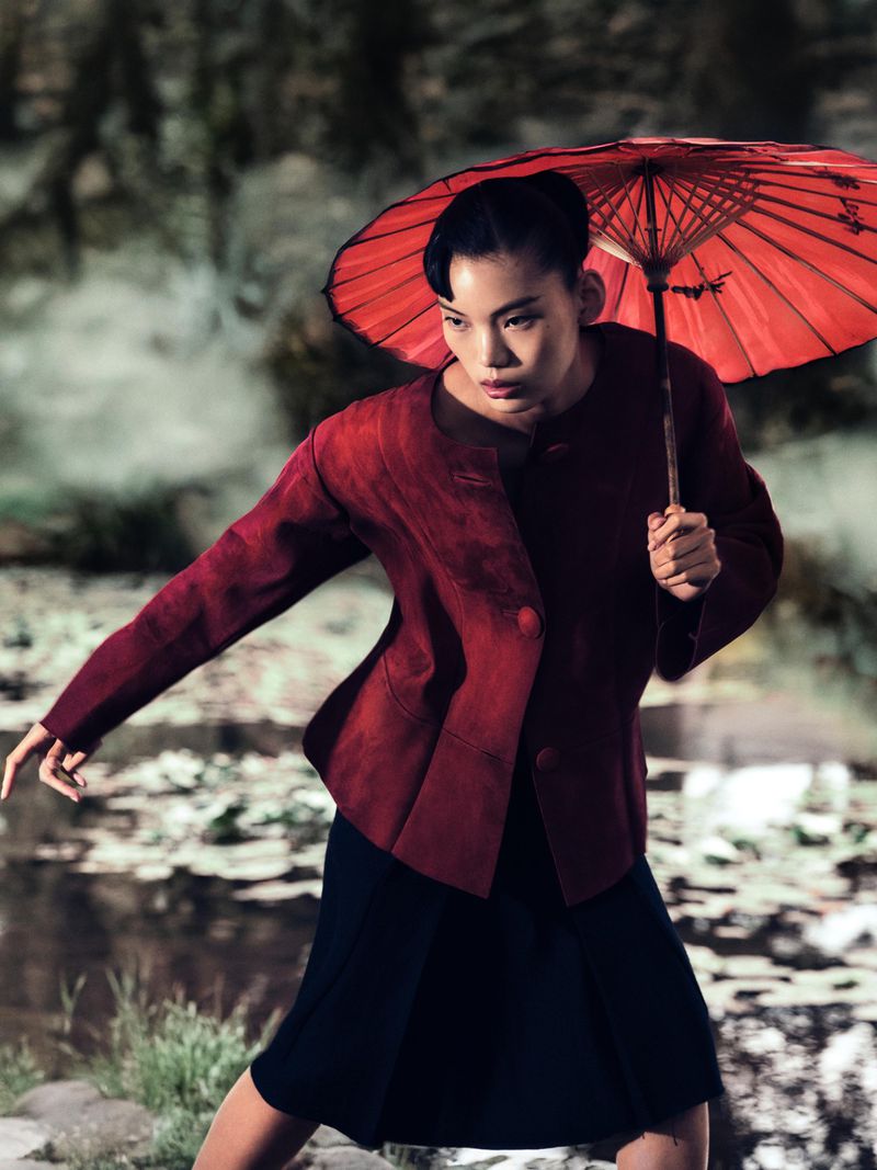 Asian woman in red jacket and black skirt poses with a red umbrella outdoors in a natural setting with water and foliage.