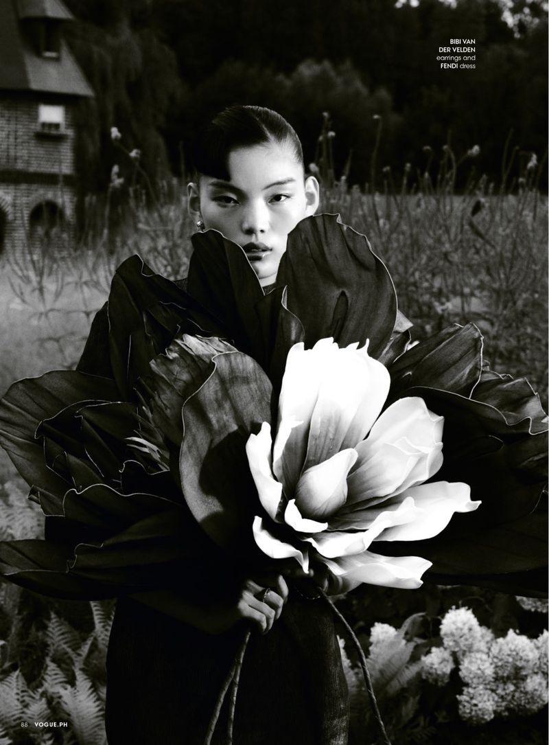 Monochrome portrait of a stylish Asian woman with slicked-back hair, holding a large flower in an outdoor setting near a brick building.