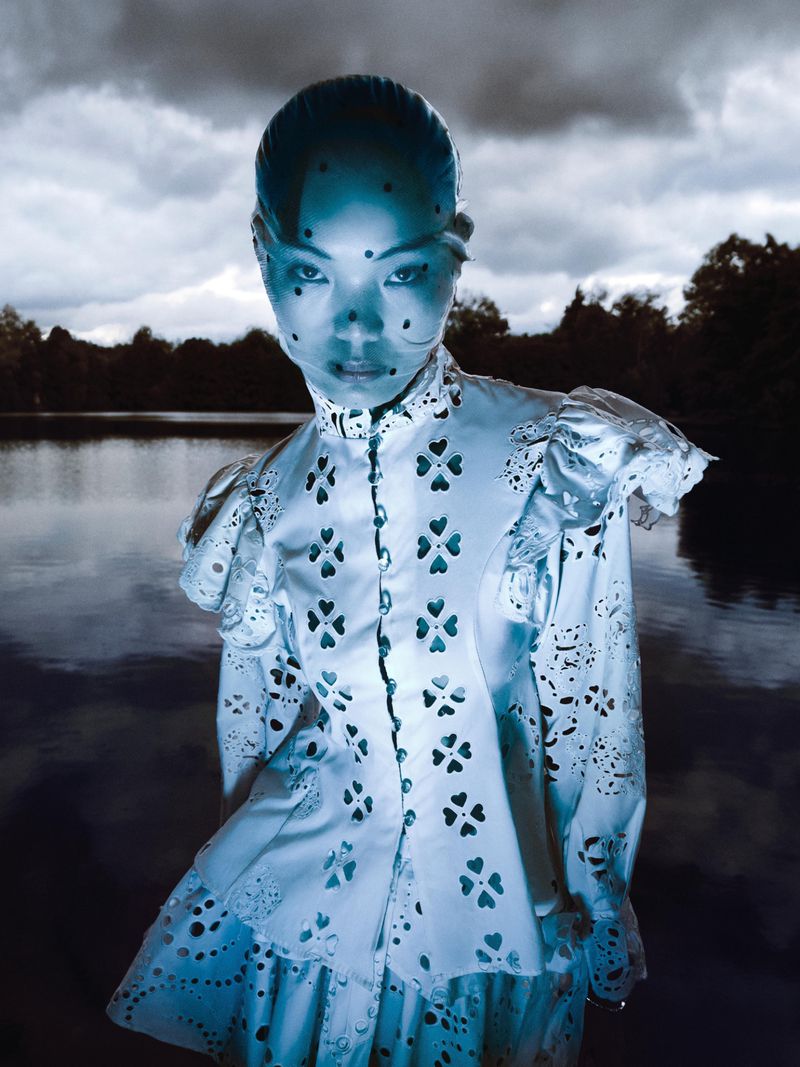 An Asian woman in a white dress with cutouts stands in front of a lake under a cloudy sky, creating a surreal and ethereal portrait.