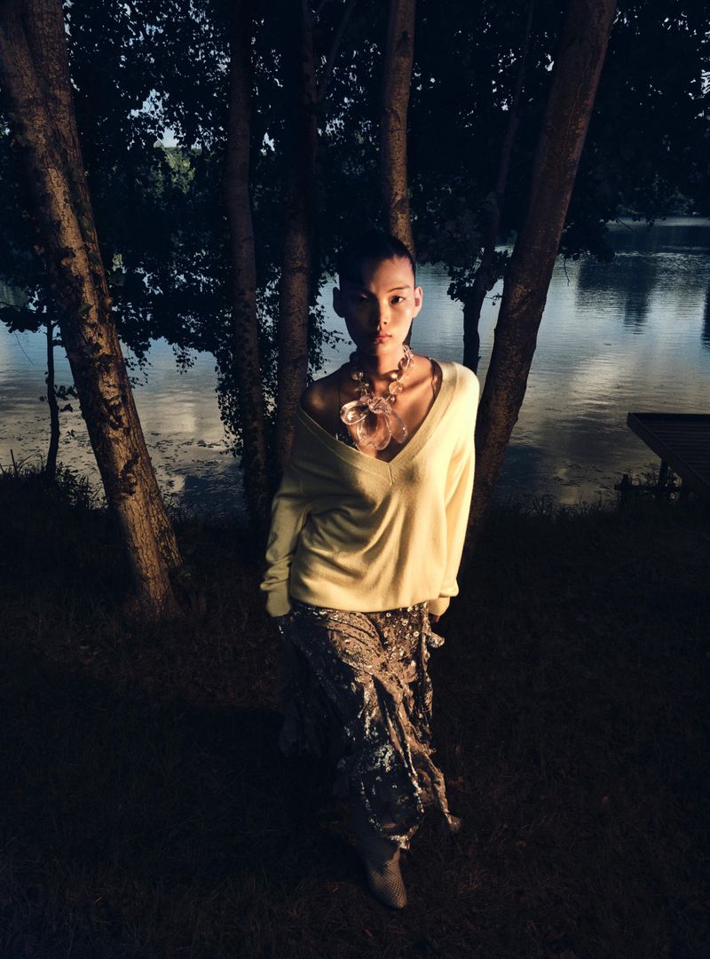 Woman in V-neck sweater and patterned skirt poses outdoors at dusk near a body of water, framed by trees, wearing statement necklace.