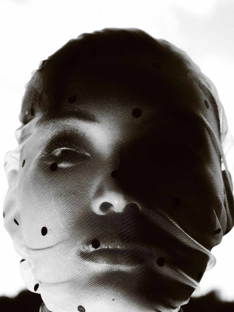 Portrait of a person with face and head covered in a dotted mesh fabric against a bright background, showcasing the mesh texture.