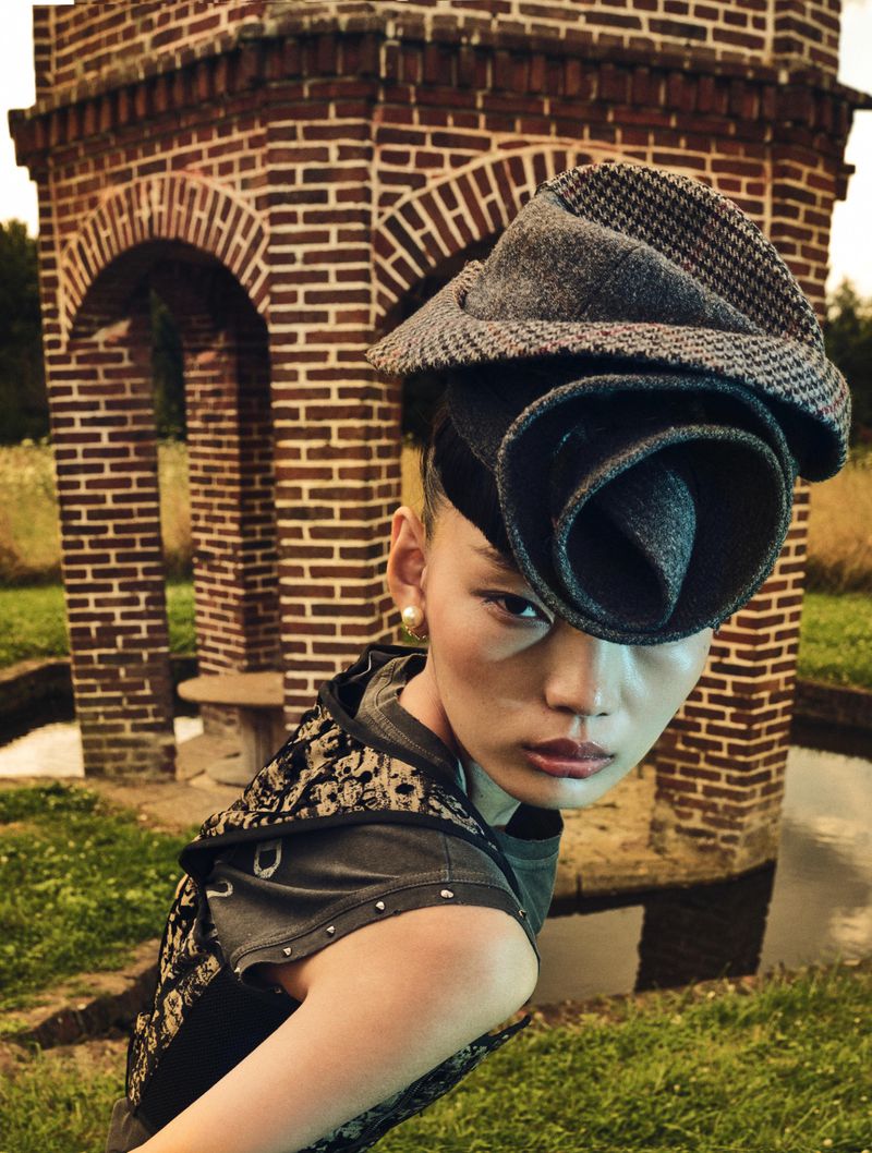 Woman poses in elaborate floral hat and patterned top outdoors, with a unique brick structure in the background on a grassy field.