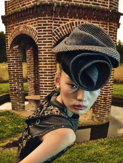 Woman poses in elaborate floral hat and patterned top outdoors, with a unique brick structure in the background on a grassy field.