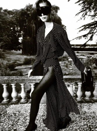 A woman in a polka-dot dress, sunglasses, and tights poses outdoors on a sunny day with trees and architecture in the background.