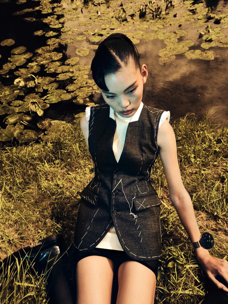 Asian woman in a dark vest poses in a grassy, outdoor setting with lily pads in the background, accessorized with a wristwatch.
