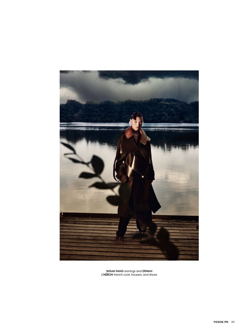 Fashion shot of adult woman in long trench coat with fur collar standing on wood dock near lake with moody landscape background.