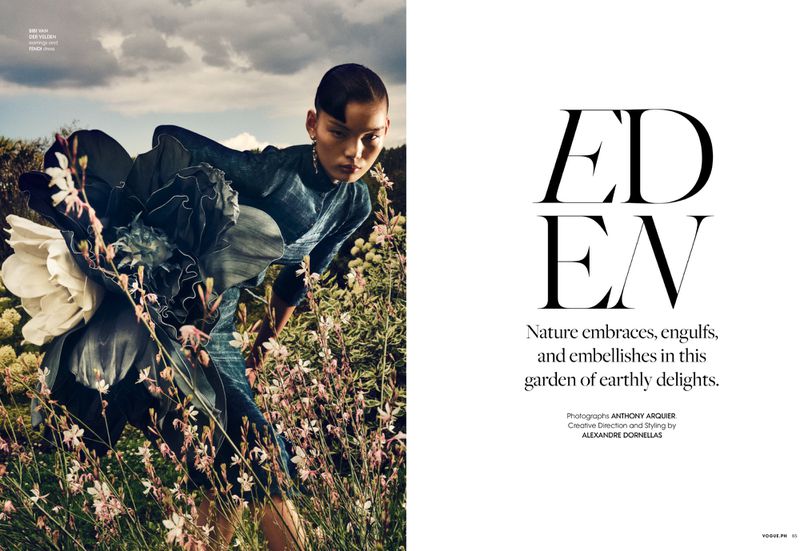Fashion shot of a slender woman in an elegant teal dress posing in a field of wildflowers under a cloudy sky, Vogue magazine.