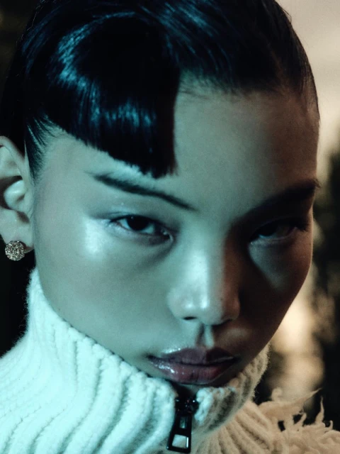 Close-up portrait of a woman with short, dark hair and fair skin, wearing a white turtleneck sweater in an outdoor setting with soft lighting.