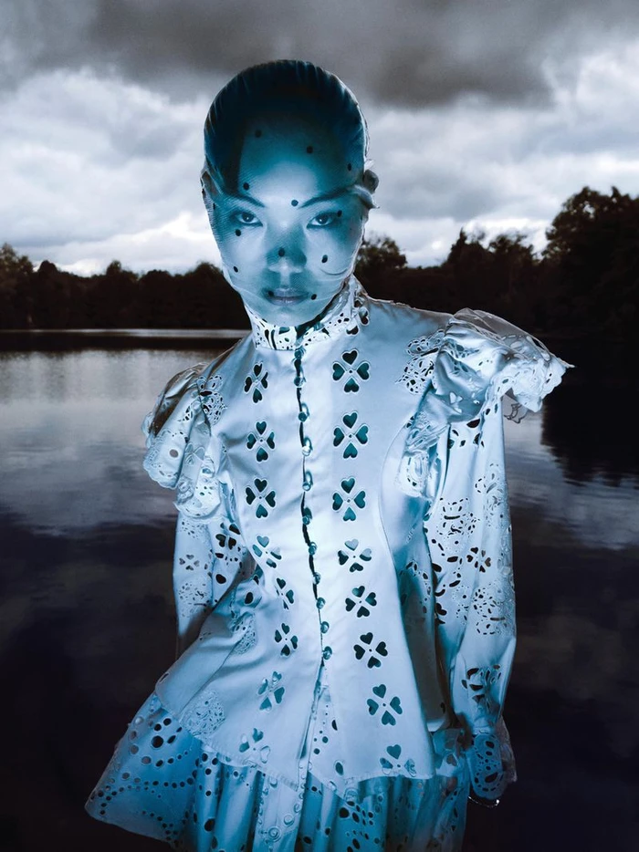 Model wearing a white cut-out patterned dress standing outdoors near a dark lake under an overcast sky.