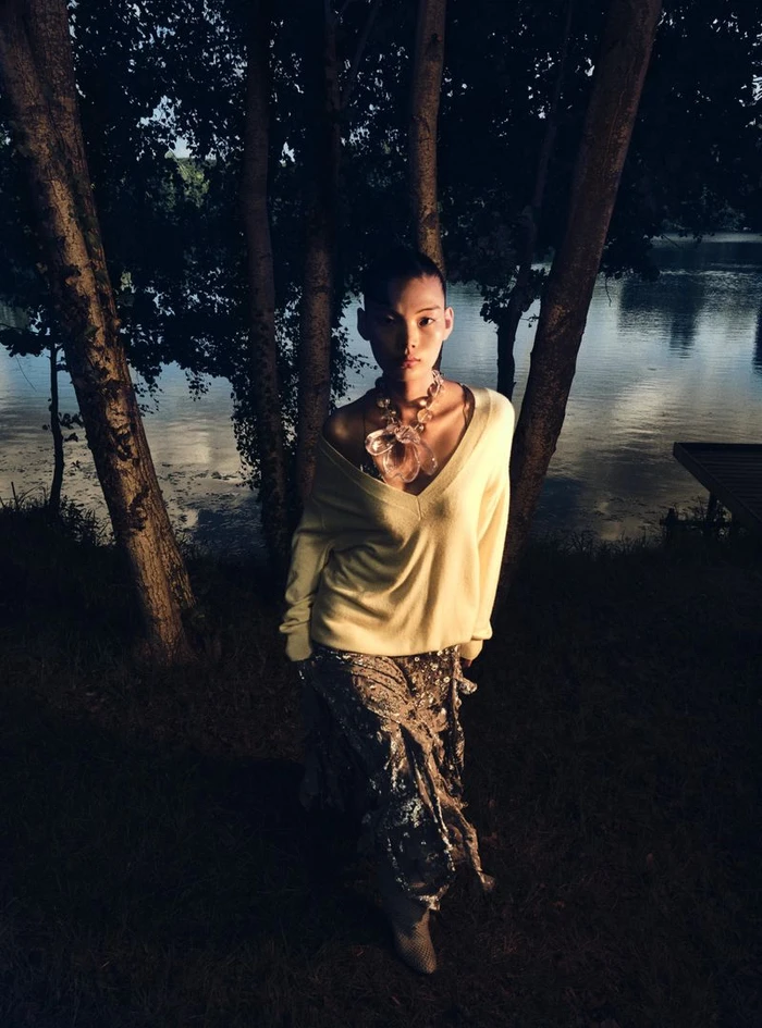 Model wearing a pale yellow V-neck sweater and patterned skirt standing among trees by a lake at dusk.