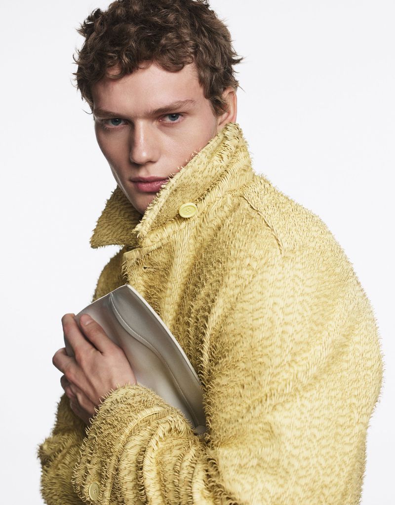 Fashion portrait of a young adult male model wearing a textured yellow coat while holding a silver clutch in a bright photo studio.