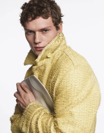 Fashion portrait of a young adult male model wearing a textured yellow coat while holding a silver clutch in a bright photo studio.
