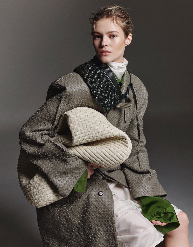 Fashion editorial shot of a model wearing a woven coat and holding a woven bag, set against a grey backdrop in a studio setting.