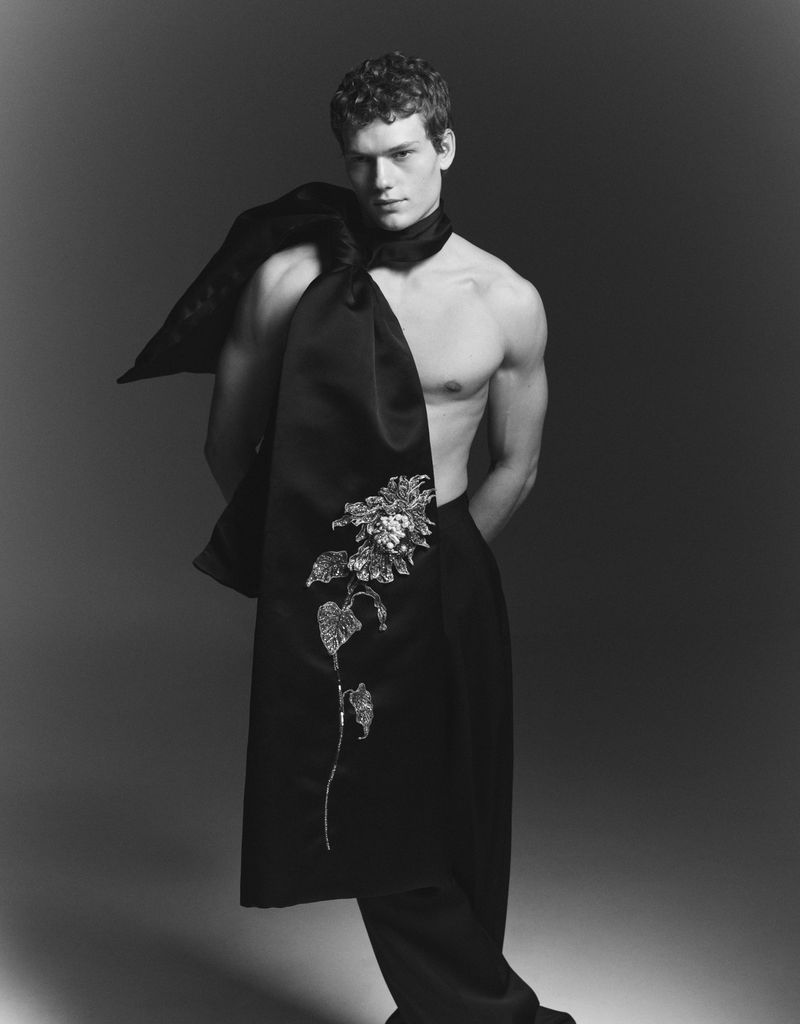 Monochrome studio shot of a shirtless male model with curly hair, wearing a black scarf with floral embroidery and draped fabric.