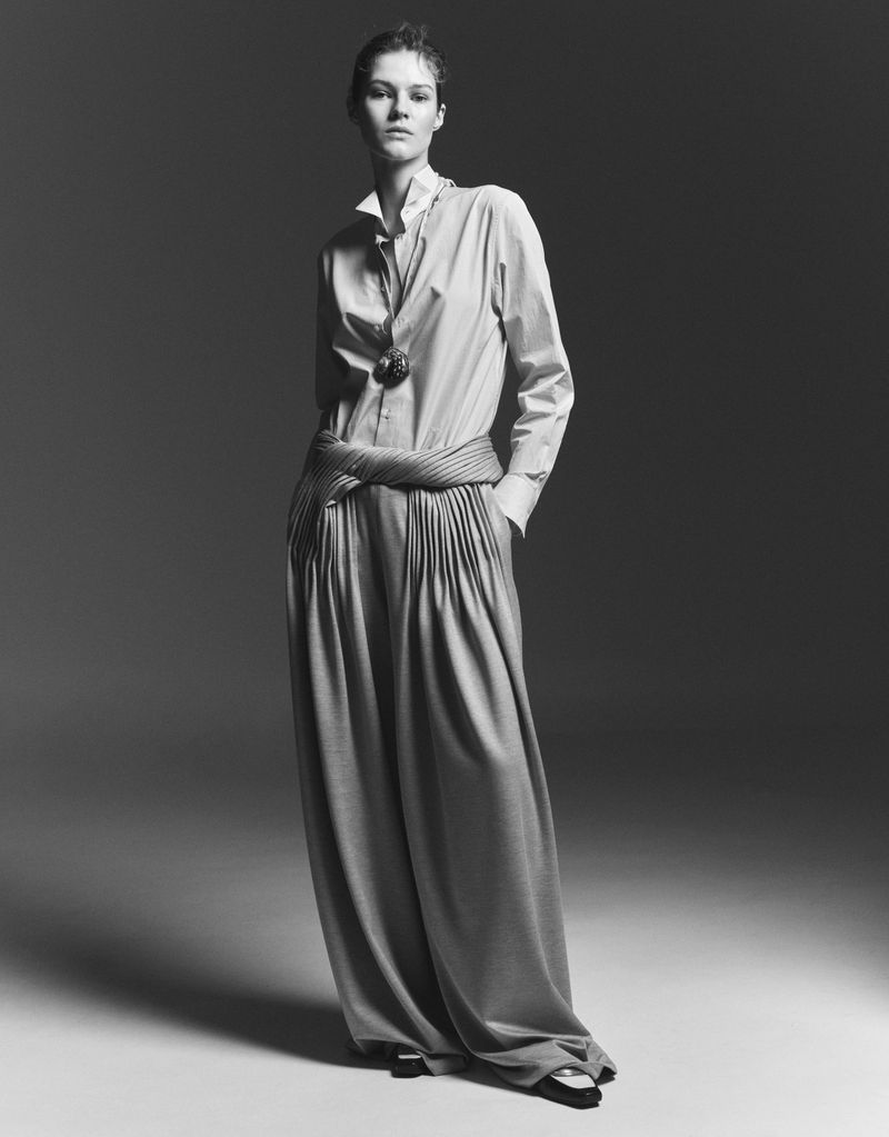 Full-length studio shot in black and white of a young woman wearing a collared shirt, wide-legged pants, and a large pendant necklace.
