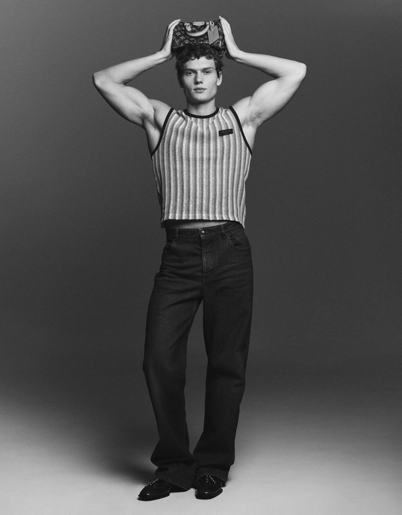 Monochromatic portrait of a male fashion model holding a handbag over his head, wearing a striped tank top and dark denim jeans.