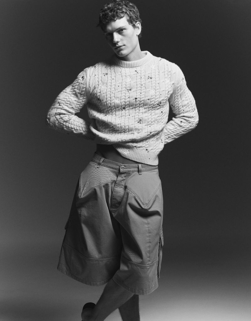 Male model wearing a textured knit sweater and baggy shorts against a dark gradient studio background.