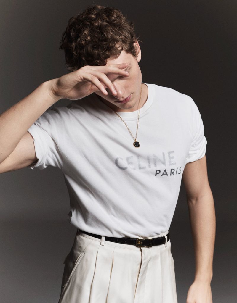 Young man in a white t-shirt with CELINE PARIS logo and beige pants, posing in a photo studio against a grey backdrop with dramatic lighting.
