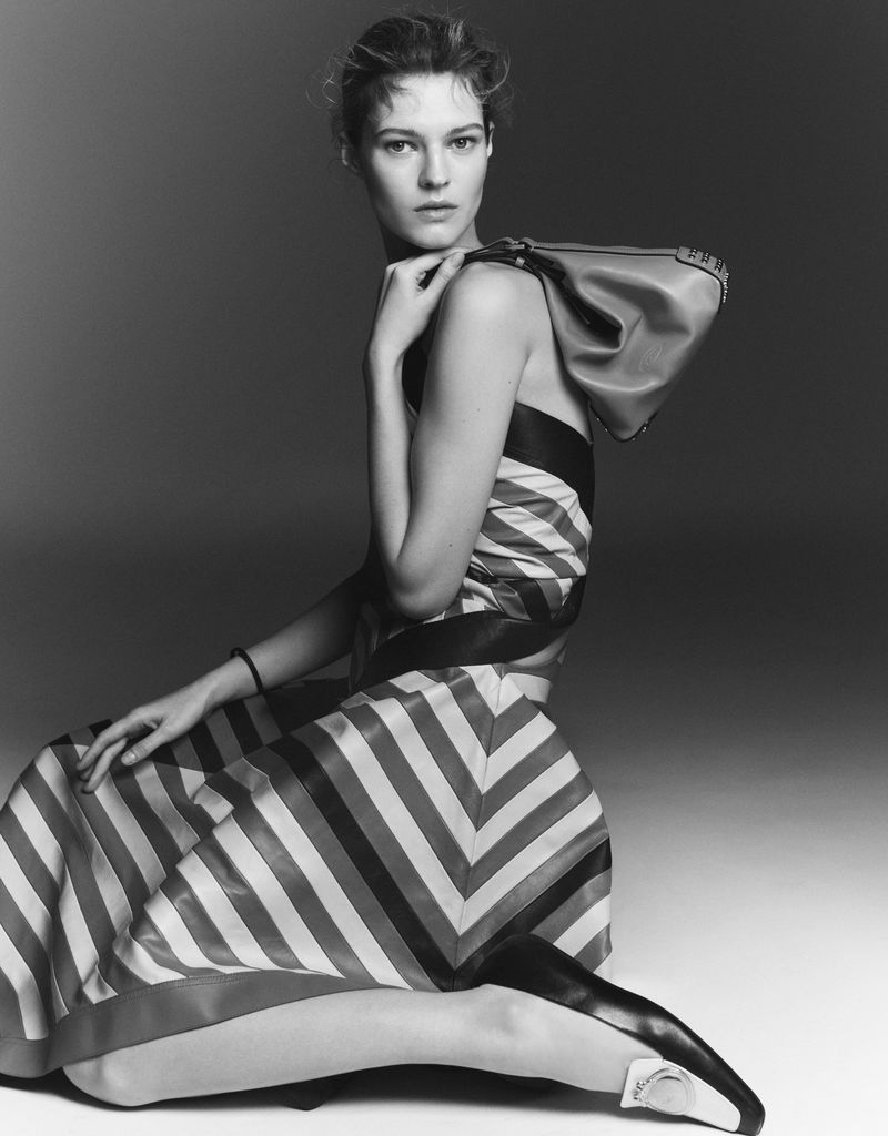 Fashion model poses in studio, kneeling while holding a handbag, wearing a striped dress and pointed-toe shoes against a neutral background.