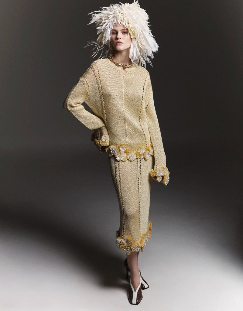 Woman in a gold knit dress with white feathered hat in a studio setting, posing in front of a neutral gray backdrop in a fashion editorial style.