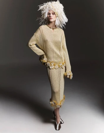 Woman in a gold knit dress with white feathered hat in a studio setting, posing in front of a neutral gray backdrop in a fashion editorial style.