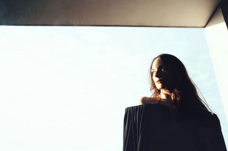 Portrait of a brunette woman in off-shoulder top, looking away against a bright sky, standing in a corner with modern architecture.