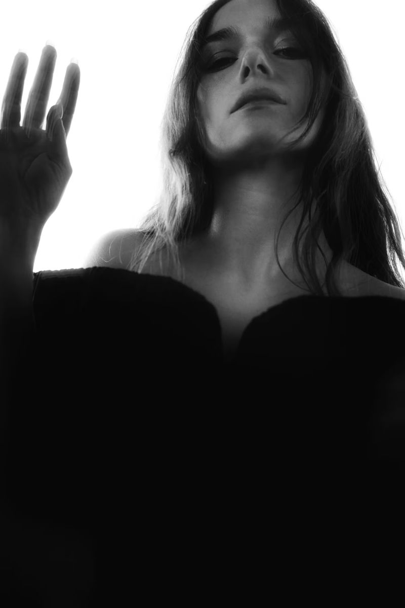 Black and white portrait of a woman looking down, hand raised, soft focus and dramatic lighting against a bright white background.