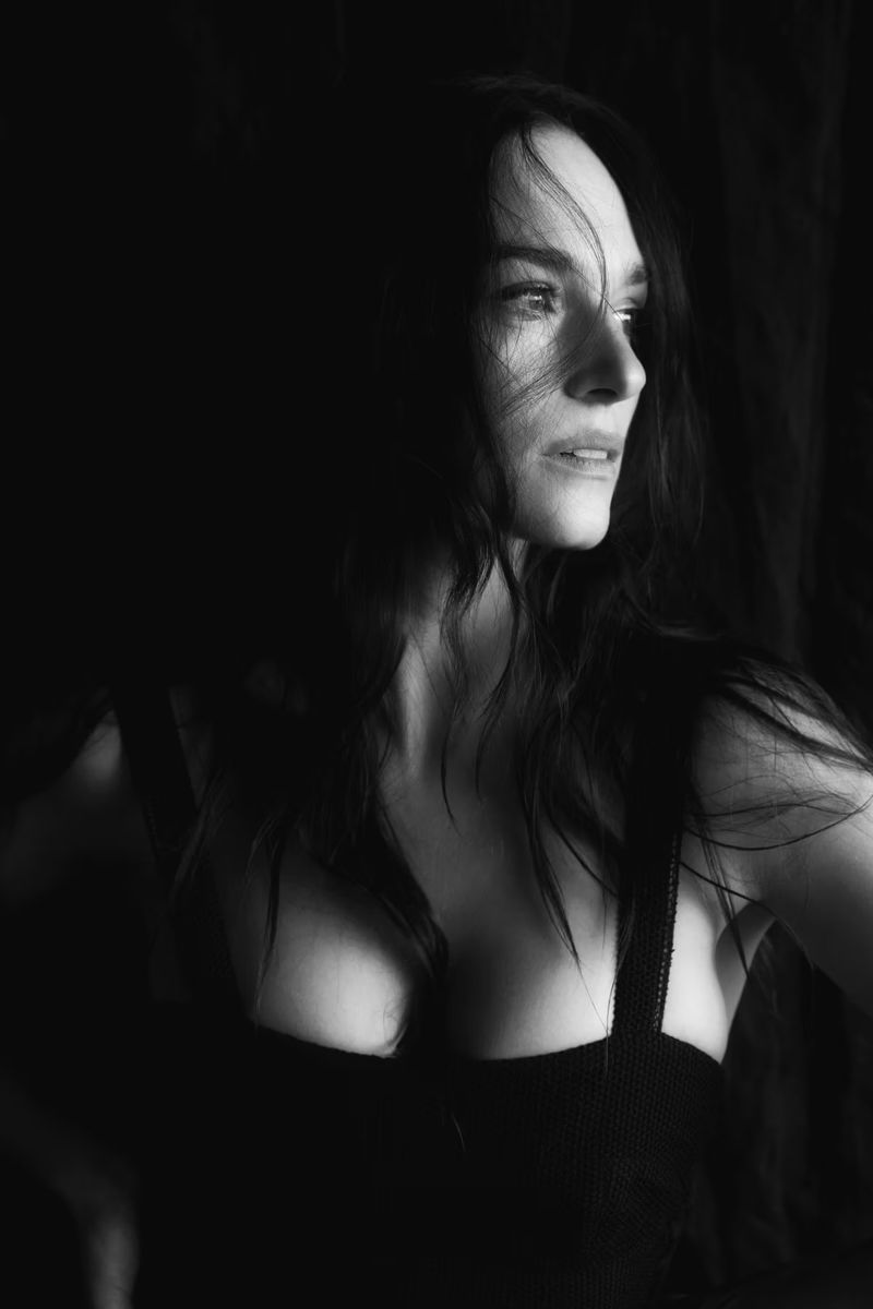 Black and white portrait of a woman with long dark hair, looking to the side in a dark indoor environment with soft, diffused lighting.