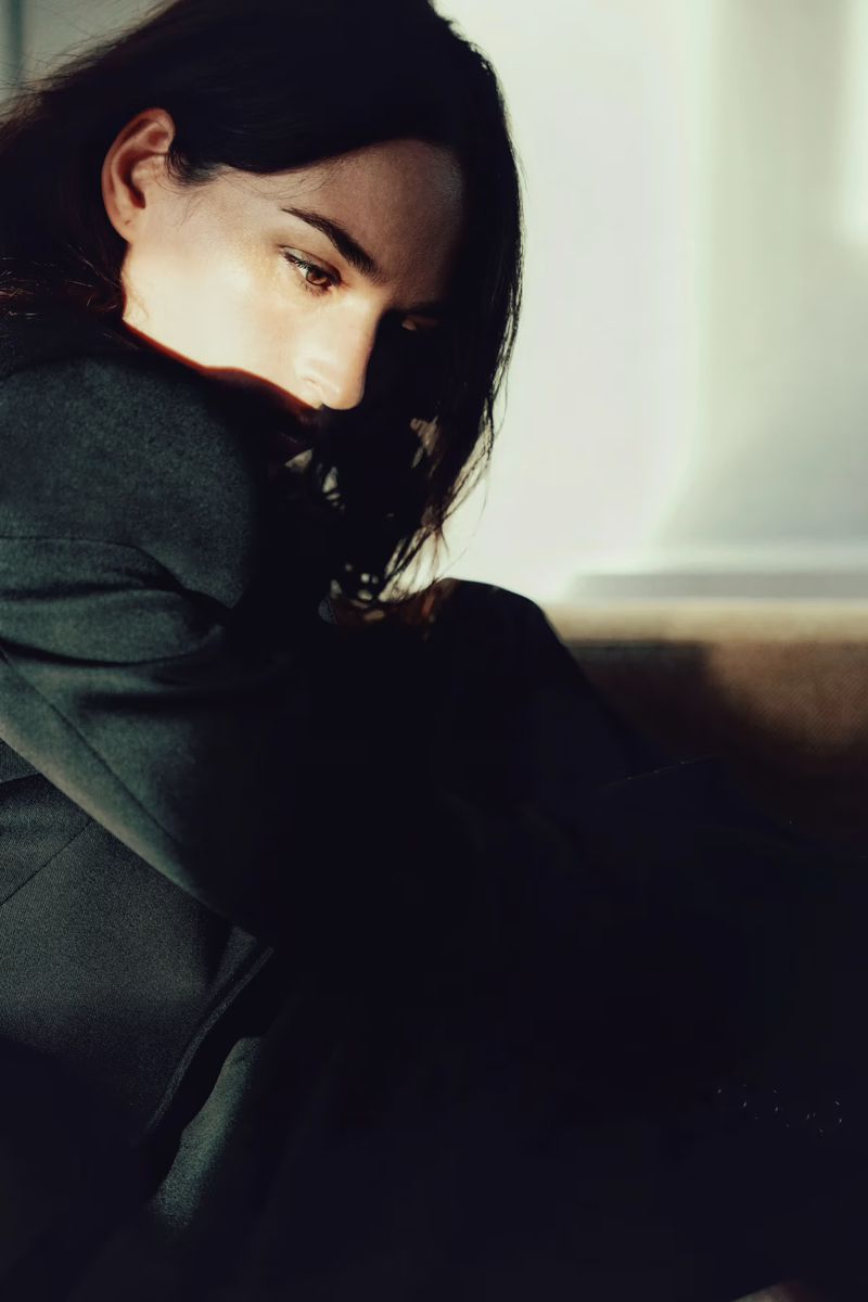 Portrait of a woman with dark hair wearing a dark blazer, partially illuminated by natural light against an interior wall.