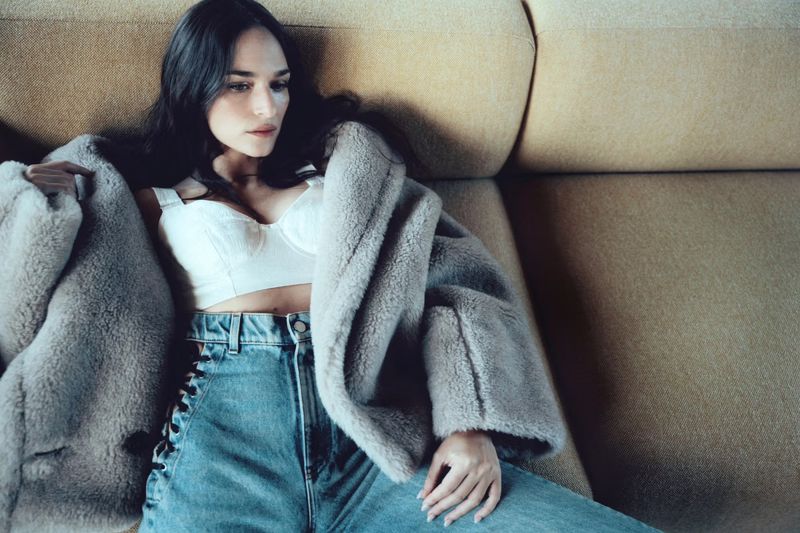 Woman relaxing on a couch, wearing jeans and a bra top, with a soft, shearling-like jacket draped over her. Indoor setting, soft natural lighting.