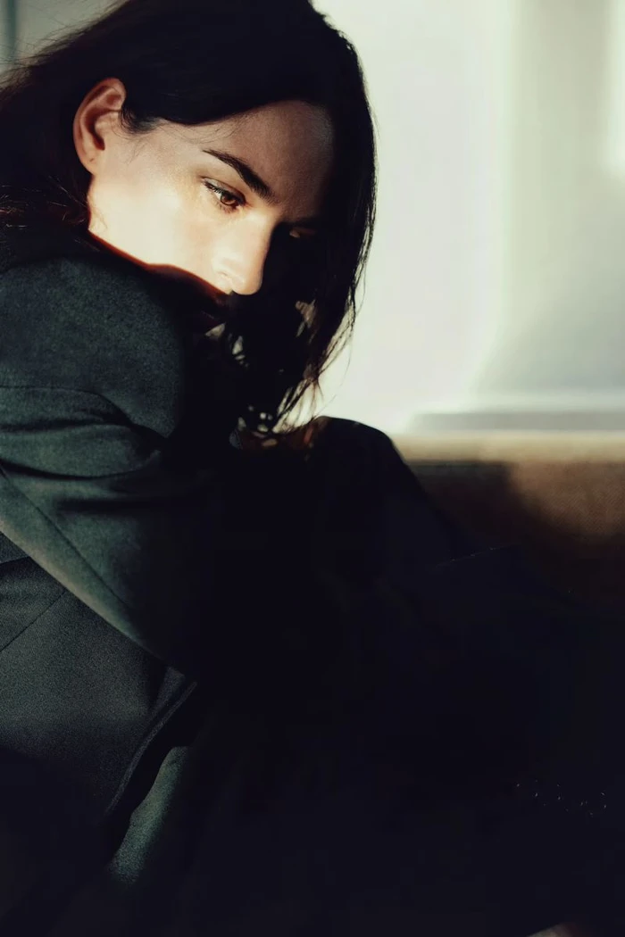Portrait of a woman with dark hair wearing a dark blazer, partially illuminated by natural light against an interior wall.