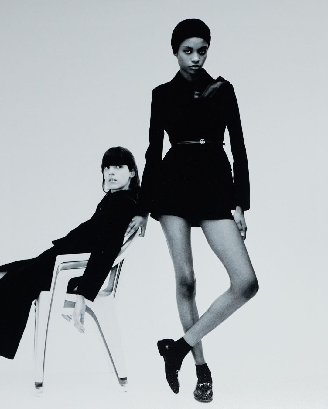 Two fashion models pose in black outfits against a neutral background, one seated on a white chair, in a high-contrast black and white studio shot.