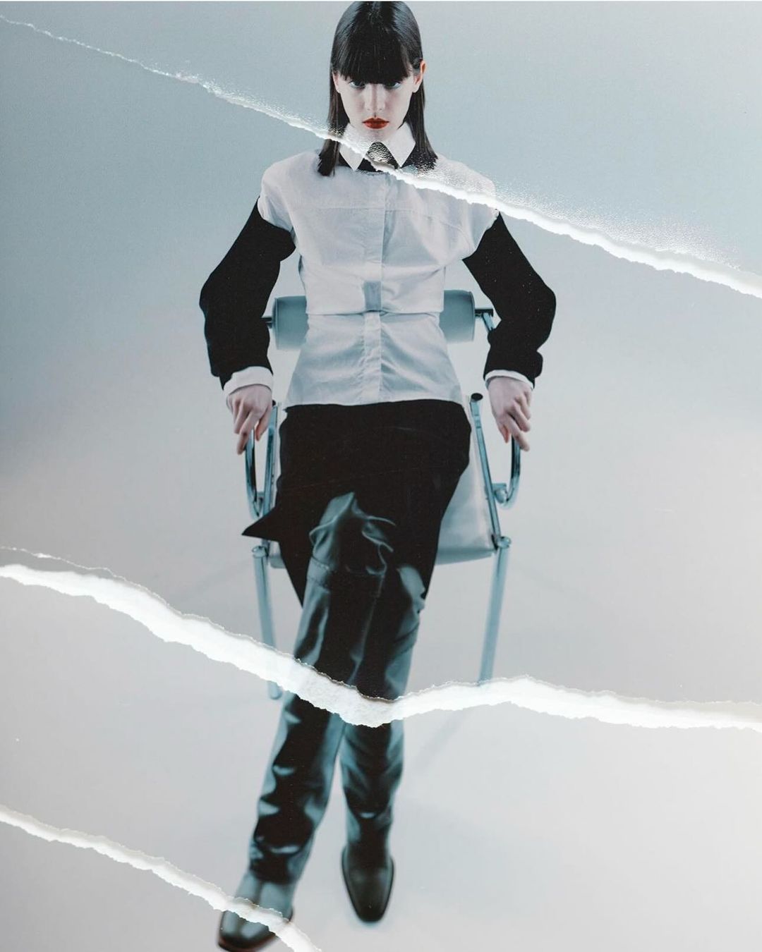 A woman sits on a metal chair in a minimalist indoor studio. She is wearing a white button-down shirt, dark pants, and leather boots.