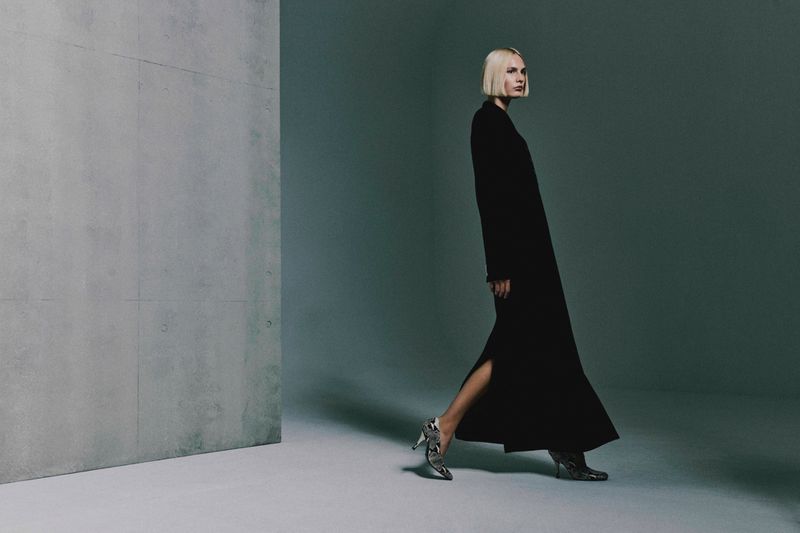 Elegant woman in a long black dress and patterned heels walks in front of a grey wall in a minimalist studio setting.