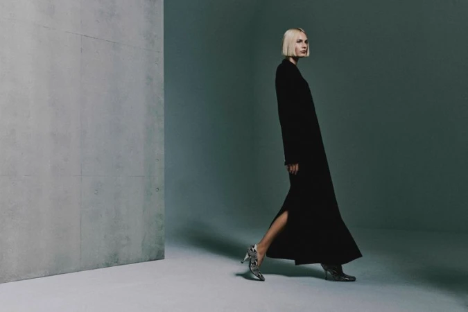 Elegant woman in a long black dress and patterned heels walks in front of a grey wall in a minimalist studio setting.
