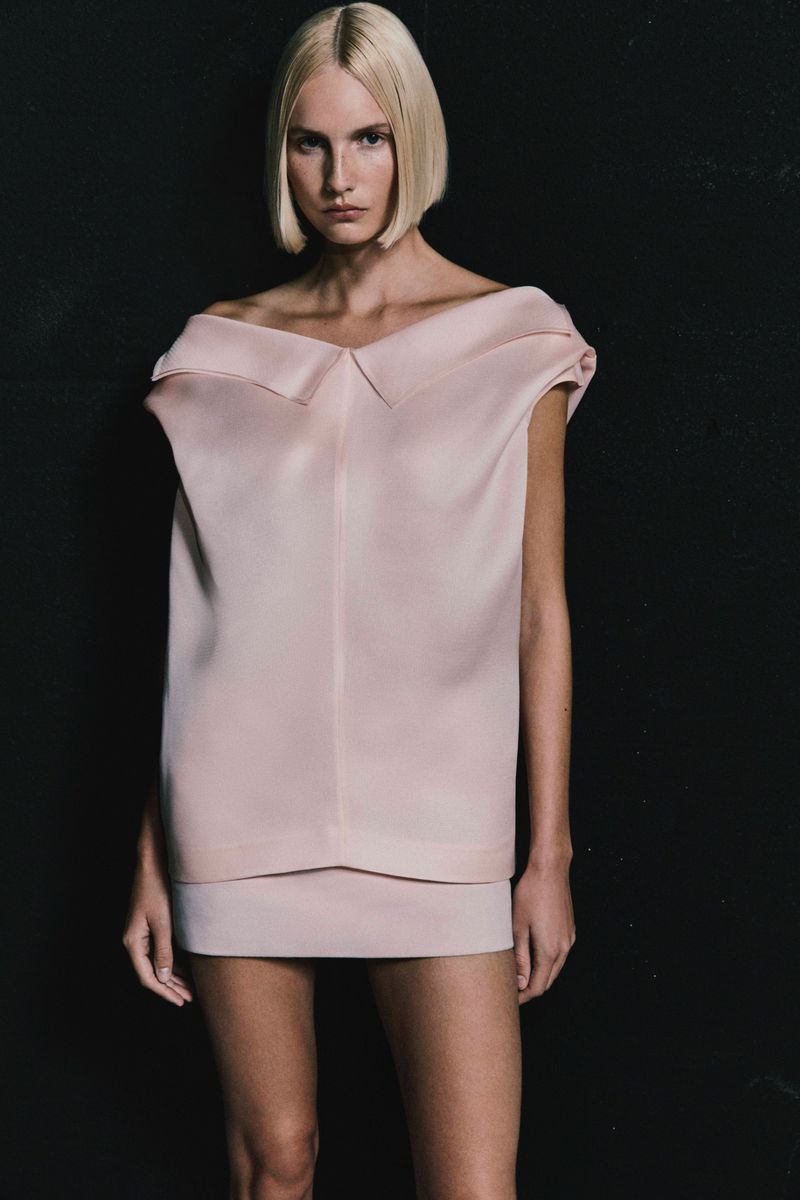 Fashion shot of a blonde adult woman posing indoors in a pink dress against a dark background, emphasizing design and style.