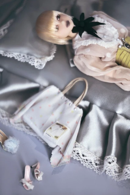Art doll in a light pink dotted dress with a black collar laying on a silver satin pillow with bags and shoes displayed.