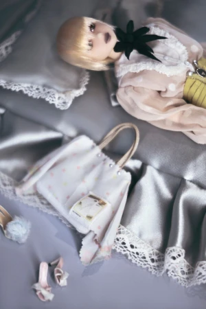 Art doll in a light pink dotted dress with a black collar laying on a silver satin pillow with bags and shoes displayed.