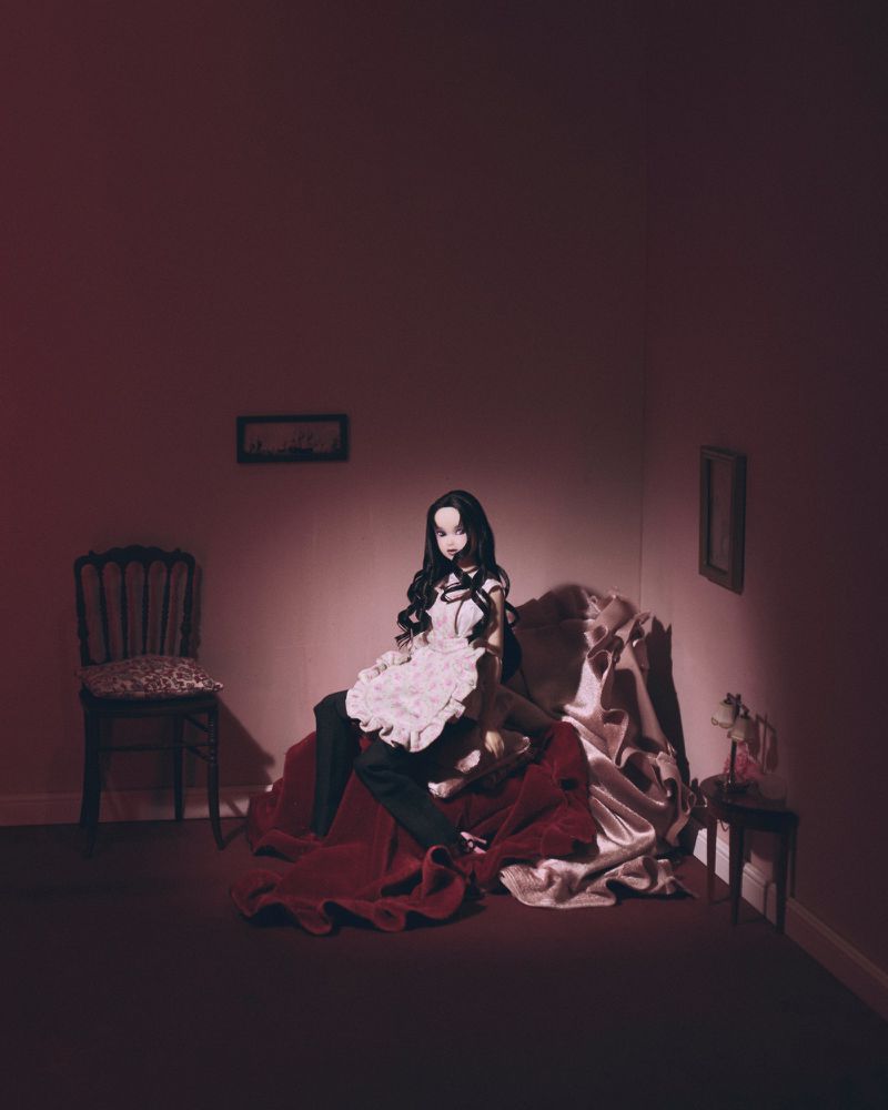 Artistic indoor photo featuring a posed fashion doll with dark hair and pale skin, wearing a white dress, seated on draped fabric.