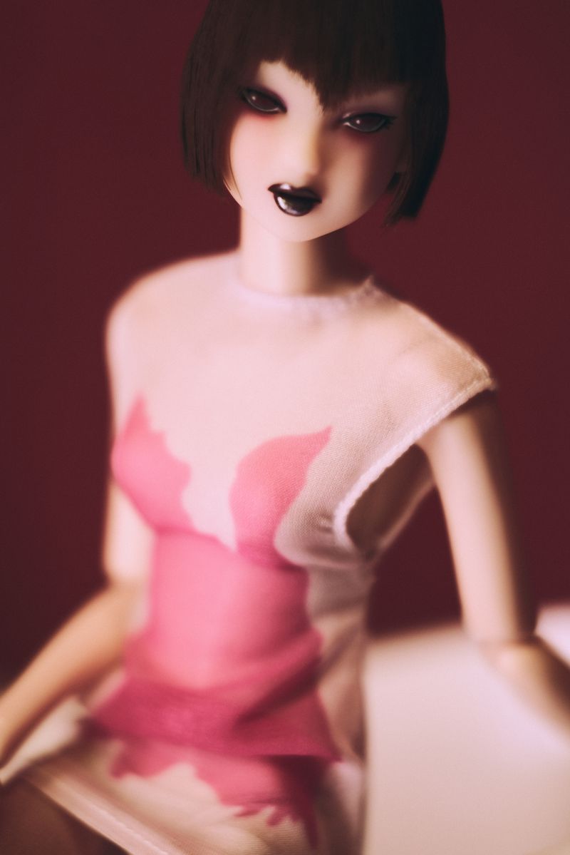A doll with pink and white dress is featured against a red background with dramatic lighting highlighting its makeup and features.