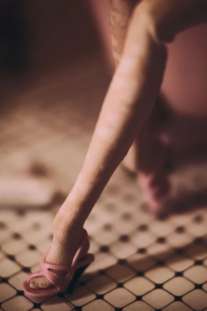 Close-up on a woman's leg and pink high-heeled sandal, set against a black and white tiled floor, conveying a vintage fashion aesthetic.