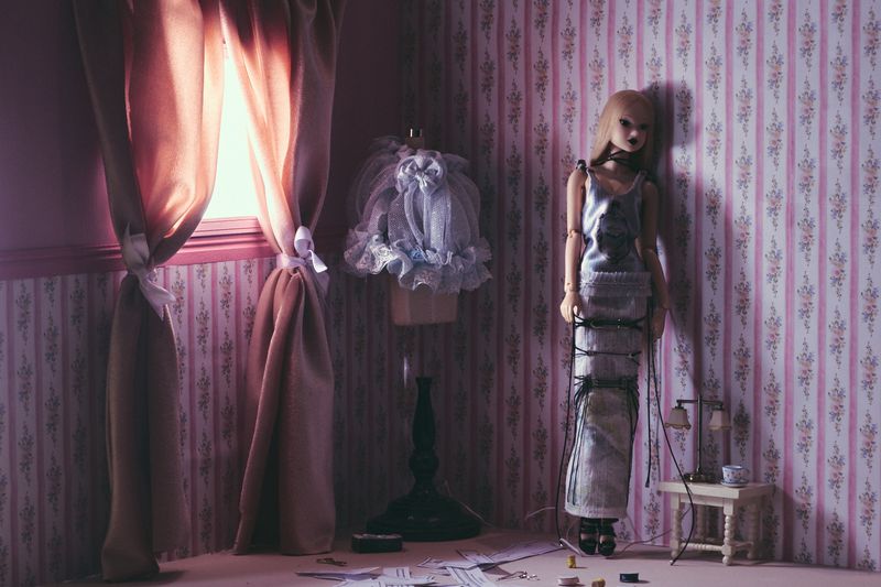 Doll in a silver dress standing in a room with floral wallpaper, next to a dress form, window with curtains, and a small table.