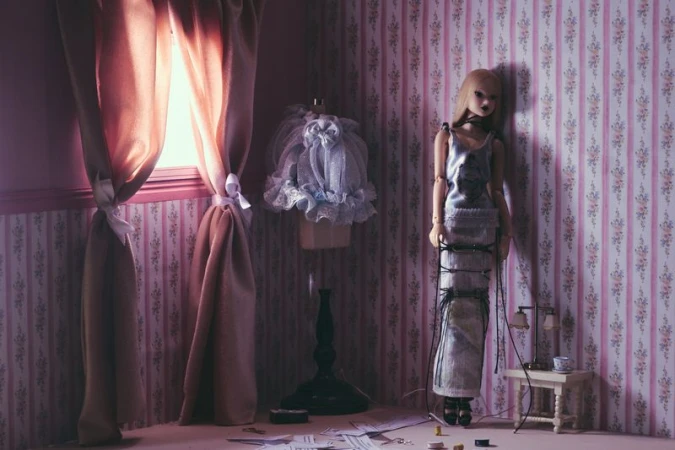 Doll in a silver dress standing in a room with floral wallpaper, next to a dress form, window with curtains, and a small table.